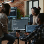 Grupo de profissionais de tecnologia colaborando em um escritório moderno, com computadores exibindo códigos e ambiente decorado com tijolos aparentes e plantas.