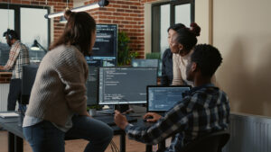 Grupo de profissionais de tecnologia colaborando em um escritório moderno, com computadores exibindo códigos e ambiente decorado com tijolos aparentes e plantas.