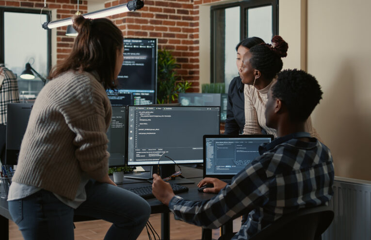 Grupo de profissionais de tecnologia colaborando em um escritório moderno, com computadores exibindo códigos e ambiente decorado com tijolos aparentes e plantas.
