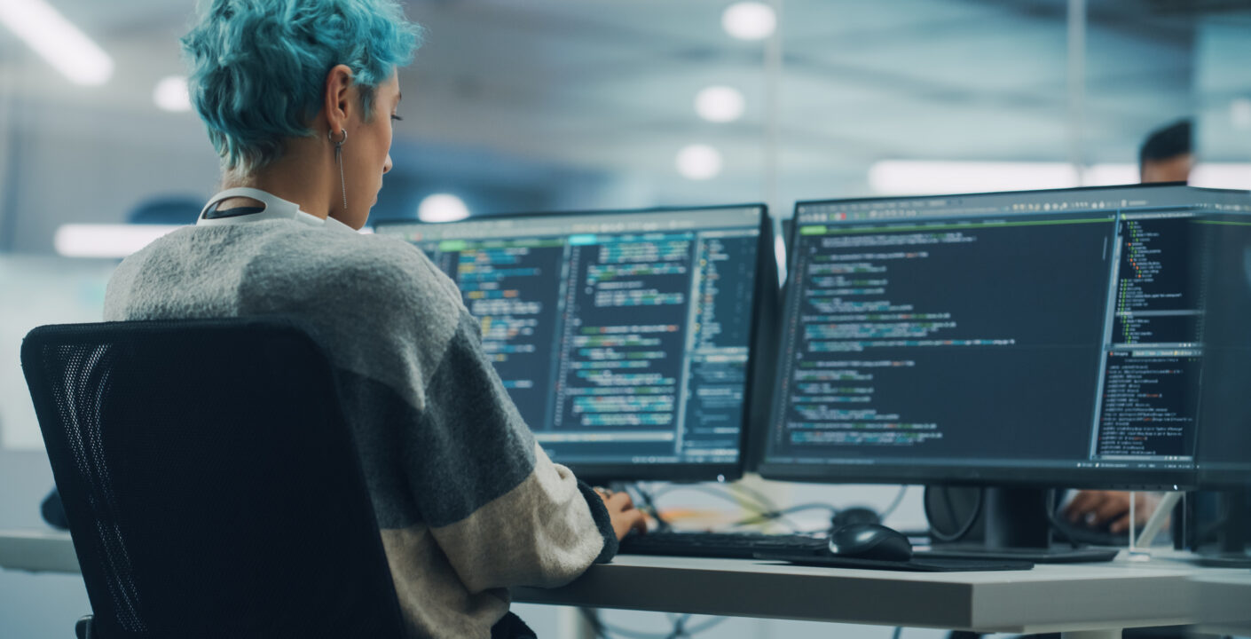 Programadora com cabelo azul trabalhando em múltiplos monitores exibindo códigos de programação, em estação de trabalho moderna no escritório.
