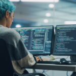 Programadora com cabelo azul trabalhando em múltiplos monitores exibindo códigos de programação, em estação de trabalho moderna no escritório.