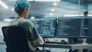 Programadora com cabelo azul trabalhando em múltiplos monitores exibindo códigos de programação, em estação de trabalho moderna no escritório.