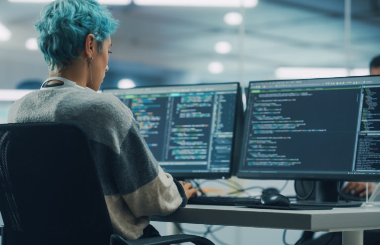 Programadora com cabelo azul trabalhando em múltiplos monitores exibindo códigos de programação, em estação de trabalho moderna no escritório.