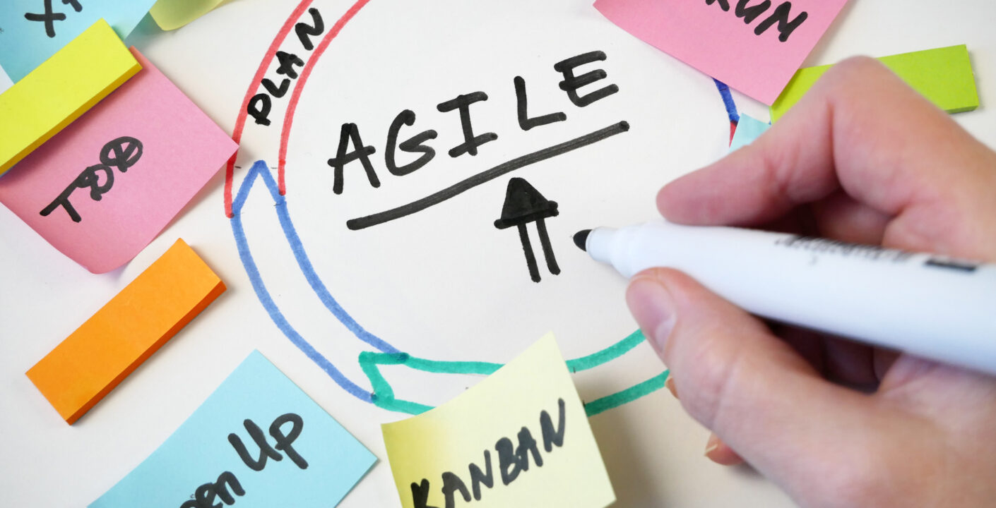 Foco na palavra "AGILE" escrita no centro de um círculo em um quadro branco, rodeada por post-its coloridos com termos como Scrum, Kanban, XP, To Do, Plan e Open Up, representando conceitos das metodologias ágeis. Uma mão segura uma caneta e destaca a palavra "AGILE".